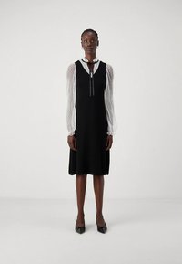 Juoda suknelė su aptemptu liemeniu ir platėjančia sijono dalimi, puošta pusiau permatomomis pūstomis taškuotomis rankovėmis ir raktelį primenančia iškirpte su kaspino akcentais.