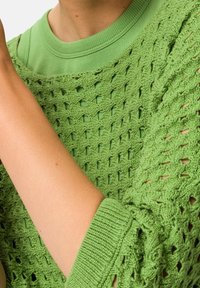 Groen gebreide trui met een opengewerkte structuur en geribbelde manchetten, gedragen over een effen groen T-shirt. Beide kledingstukken hebben een textuur.