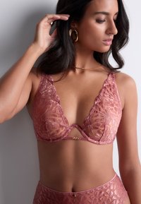 Bralette en dentelle de couleur rose doux avec des bords festonnés et une découpe centrale. Présente des motifs floraux complexes et des accents en matériel doré.