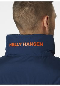 Chaqueta acolchada azul marino con cuello alto, que presenta el nombre de la marca "HELly HANSEN" en texto naranja brillante en la parte posterior. Textura suave.