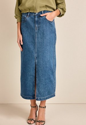 Jupe midi en denim bleu avec fente avant, portée avec une veste vert olive et des sandales à talons hauts en cuir imprimé serpent, main dans la poche.
