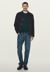 Giacca blu con bottoni sopra un maglione verde, jeans blu e scarpe nere. L'outfit presenta un design casual con tessuti testurizzati.