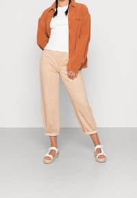 Rostfarbene Cordjacke über einem weißen, gewellten Tanktop, kombiniert mit hellbeigen, lockeren Hosen und weißen Keilsandalen.