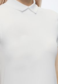 Personne portant un polo blanc uni à manches courtes avec un col plat, visible des épaules jusqu'à la partie supérieure de la poitrine et du cou.