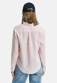 Camicia button-up rosa chiaro con strisce verticali bianche, maniche lunghe e orlo arrotondato. Abbinata a jeans in denim blu e un cappellino blu navy.