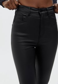 Schwarze Kunstleder-Hosen mit hohem Bund, Fronttaschen und sichtbaren Steppnähten. Glatte Textur und schlankes Design.