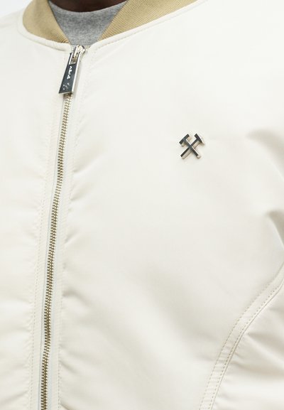 Jachetă bomber crem cu guler ribbed bej, detaliată cu un fermoar argintiu și un logo din metal negru pe piept. Textură netedă.