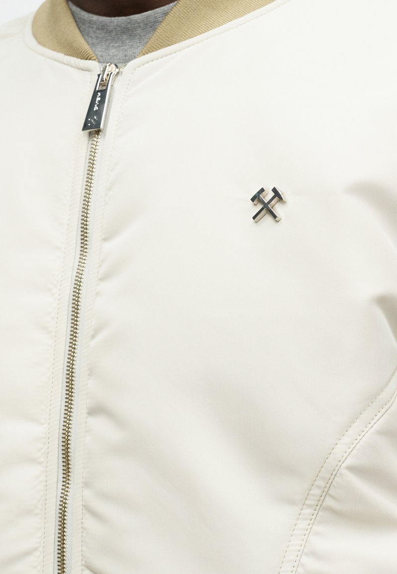 Crème bomberjack met een beige geribbelde kraag, afgewerkt met een zilveren rits en een zwart metalen logo op de borst. Soepele textuur.