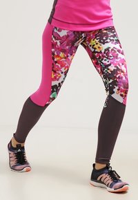 Piernas en leggings florales coloridos y rosas con zapatillas deportivas negras y rosas en una postura amplia sobre un suelo blanco.