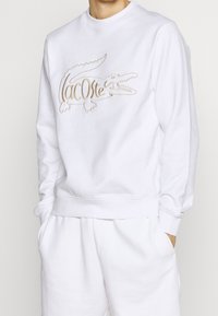 Sweat-shirt en coton blanc avec un grand logo crocodile en relief de couleur beige. Il possède un col rond ainsi que des poignets et un ourlet côtelés.