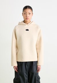 Beige kapucīna džemperis ar melnu logo, kuram ir brīvs piegriezums, raglanu piedurknes un gluda tekstūra. Apvienots ar melniem biksēm.