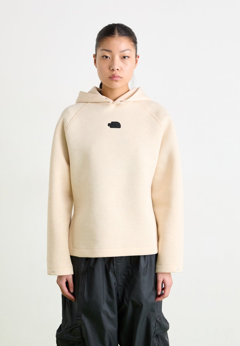 Beige kapucīna džemperis ar melnu logo, kuram ir brīvs piegriezums, raglanu piedurknes un gluda tekstūra. Apvienots ar melniem biksēm.