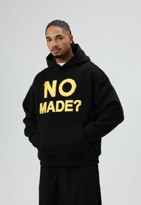 Zwarte hoodie van fleece materiaal, met gedurfde gouden letters "NO MADE?" op de voorkant. Voorzien van een voorzak en geribbelde manchetten.