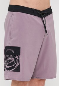 Lilla svømmeshortser med svart midjebånd og en sideficka som har et svart grafisk design. Materialet ser lett og glatt ut.