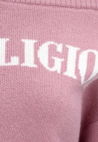 Pull rose tricoté avec de grandes lettres blanches. Tissu texturé avec encolure côtelée, montrant une légère peluche et douceur.