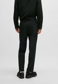Schwarze strukturierte Hose mit passgenauer Form, flachem Bund und zwei Gesäßtaschen. Getragen mit einem langärmeligen schwarzen Hemd und klobigen schwarzen Schuhen.