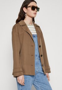 Bruine blazer jas met grote kraag, knoopsluiting en zijzakken. Gecombineerd met een gestreepte top en spijkeroveralls. Zwarte zonnebril en hartvormige oorbellen.