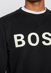 Persona que lleva una sudadera negra con grandes letras blancas "BOS" visibles en el frente y un cuello blanco asomando por el cuello.