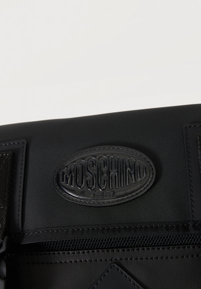 Μαύρη τσάντα με ραμμένη οβάλ δερμάτινη ετικέτα με ανάγλυφο το όνομα της μάρκας "MOSCHINO" και διακοσμητικό πάνω ράψιμο.