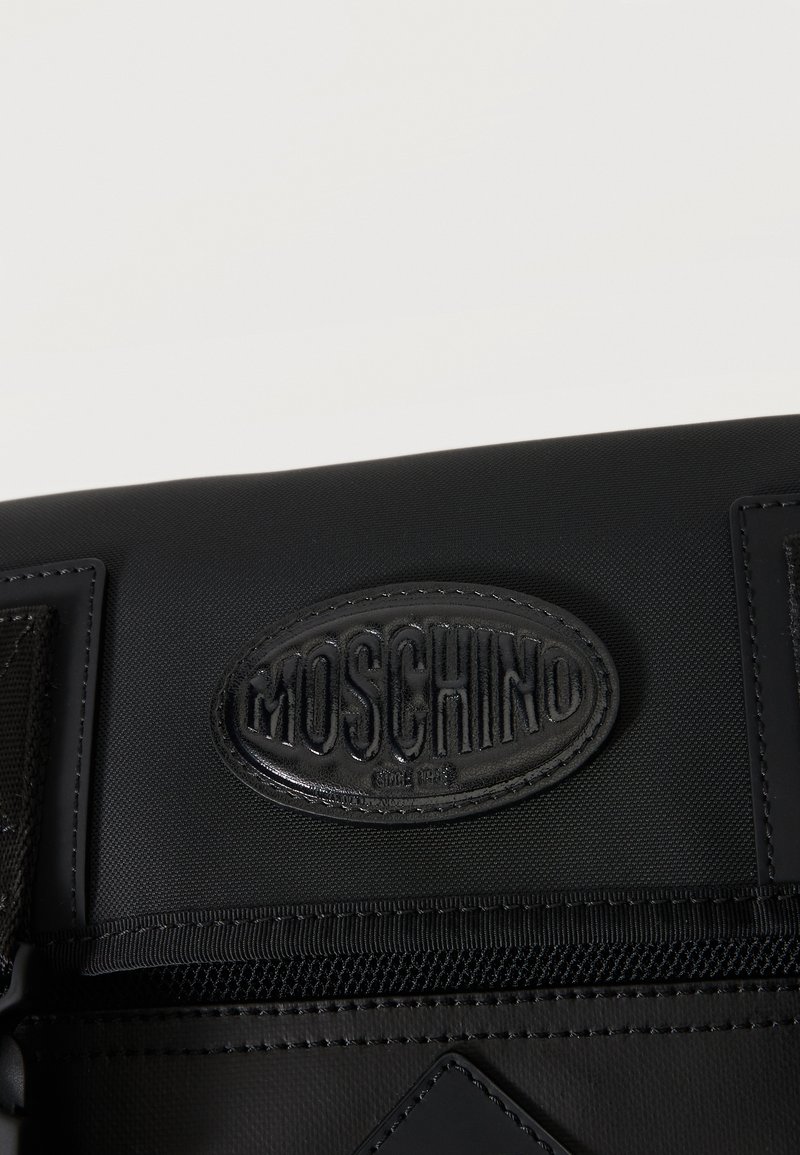 Sac noir avec un patch ovale en cuir cousu, embossé du nom de la marque "MOSCHINO" et une surpiqûre décorative sur le dessus.