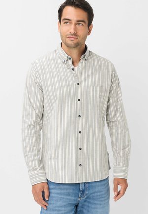 Mann trägt ein hellgraues und weißes, vertikal gestreiftes Hemd und blaue Jeans, steht vor einem schlichten hellen Hintergrund.