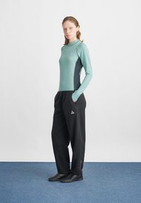Bluză verde deschis cu mânecă lungă, cu panouri laterale întunecate, asociată cu pantaloni de trening negri. Topul are un guler înalt și un logo mic. Încălțăminte neagră.