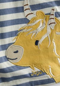 Vyšívaný design žltého vysokohorského býka na modro-bielom prúžkovanom materiáli. Obsahuje textúrované stehy a malú etiketu "JoJo".