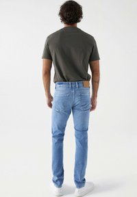 Jeans de mezclilla azul claro con corte ajustado, cinco bolsillos y una parche de cuero marrón en la parte posterior de la cintura, llevados con una camiseta verde oscura.