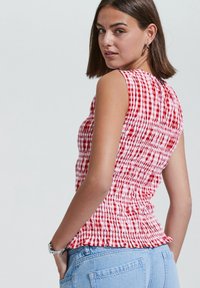 Red and white checkered sleeveless top with ruched texture, a round neckline, and a ruffled hem. Styled with light blue denim shorts.
