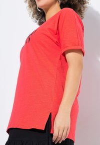 T-shirt rouge à manches courtes avec une coupe décontractée, présentant des fentes sur les côtés et une texture douce. Associé à une jupe noire en couches en bas.