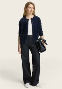 Marinefarbener, flauschiger Cardigan mit Rundhalsausschnitt und Knopfverschluss, getragen über einem weißen Oberteil und dunkelblauen weiten Jeans, kombiniert mit einer schwarzen Handtasche.