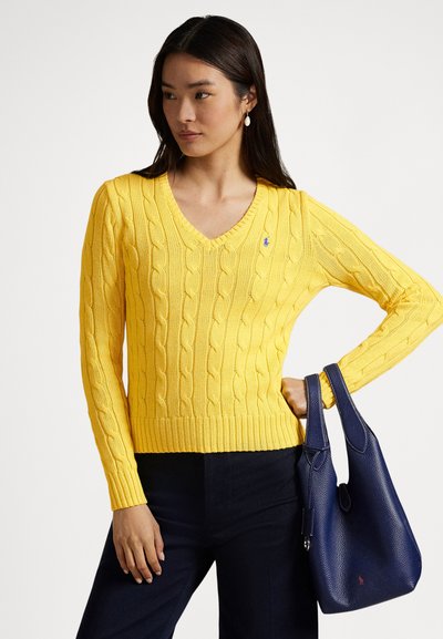 Suéter amarillo de tejido de punto con cuello en V, dobladillo acanalado y pequeño logo. Combinado con un bolso de cuero azul marino. La tela tiene una apariencia texturizada.
