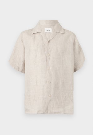Homme debout, les mains dans les poches, portant une chemise beige à manches courtes boutonnée, un short bleu marine et des sandales noires sur fond blanc.