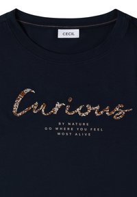 T-shirt bleu marine avec texte étincelant "Curious" et texte plus petit en dessous disant "BY NATURE GO WHERE YOU FEEL MOST ALIVE", étiquette de la marque "CECIL".