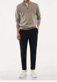 Maglione beige chiaro con zip a un quarto e maniche lunghe, abbinato a pantaloni neri slim-fit e sneakers bianche. Design minimalista, texture liscia.