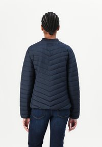 Marineblå puffjakke med chevron-søm og rund krage, med en glatt tekstur og ribbede mansjetter. Brukt med mørke jeans.