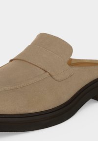 Μπεζ μοκασίνι από καστόρι slip-on με φαρδιά λωρίδα πάνω και μαύρη καουτσούκ σόλα, εμφανίζεται σε άσπρο φόντο.