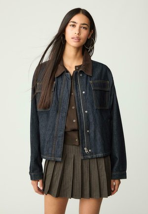 Dark denim jacket with brown corduroy collar, featuring two chest pockets, front zipper, and contrast stitching, worn over a brown top.