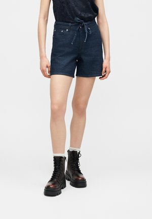 Femme portant un short en jean foncé avec une ceinture à nouer, des bottes de combat noires à lacets et des chaussettes blanches à bords festonnés, debout devant un fond uni.