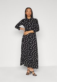 Robe maxi noire à pois avec des manches longues, un col rabattu, une fermeture à boutons sur le devant et un ourlet fluide. Portée avec des talons noirs à lanières.