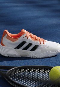 Λευκό και πορτοκαλί παπούτσι τένις Adidas δίπλα σε ρακέτα και μπάλα τένις σε μπλε επιφάνεια γηπέδου.