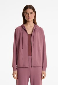 Helle lila Zip-Hoodie mit Kapuze, ausgestattet mit Raglanärmeln, Fronttaschen und strukturiertem Stoff. Getragen über einem passenden Oberteil.