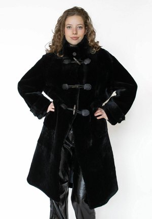 Ung kvinde med krøllet hår iført en lang sort faux pelsjakke med knapper og sorte skinnende bukser, hænderne på hofterne.