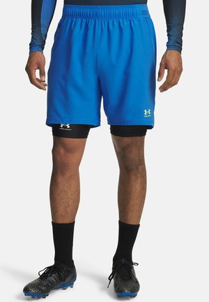 Pantalones cortos deportivos azules con una cinturilla elástica, con logos de Under Armour. Llevados sobre pantalones cortos de compresión negros y combinados con botas de fútbol negras.