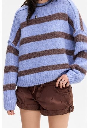 Woman wearing a light blue and brown striped fuzzy sweater and brown shorts, with rings and a pendant necklace visible.