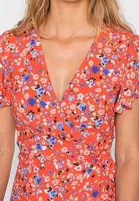 Robe portefeuille fleurie avec un profond décolleté en V et de courtes manches volantes. Caractérisée par un fond rouge orné de motifs floraux bleus, roses et blancs. Tissu lisse.