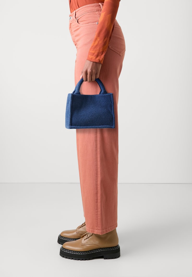 Blå filt totebag med korte håndtak, rektangulær form og kontrasterende kanter. Brukt med lyserøde bukser og brune snørebåndsankelstøvler.