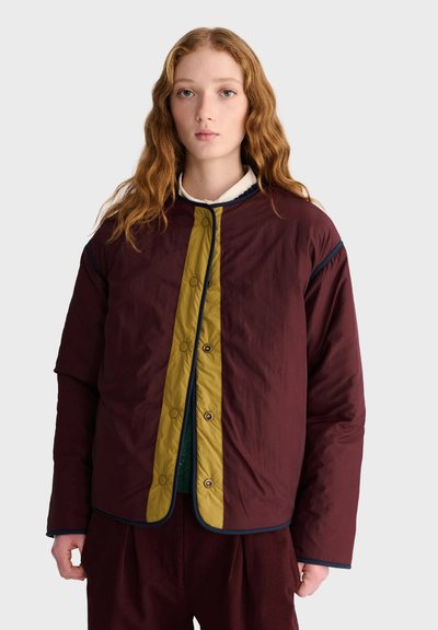 Jeune personne aux longs cheveux roux ondulés portant une veste matelassée bordeaux et olive, debout devant un fond uni.