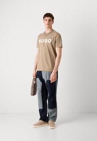 Beige bavlněné tričko s logem "HUGO", v kombinaci s barevně blokovanými denimovými kalhotami v modrých odstínech a béžovými teniskami. Drží malou crossbody tašku.
