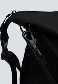 Dettagli di uno zaino nero con un gancio in metallo, una tracolla textured e un tessuto liscio. Cuciture visibili e hardware regolabile.
